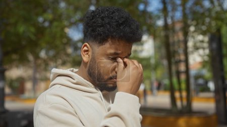 Foto de Hombre afroamericano estresado pellizcando puente de nariz en el entorno del parque de la ciudad al aire libre. - Imagen libre de derechos