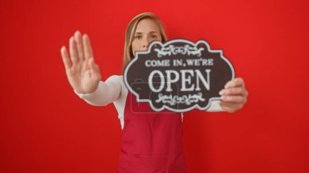Photo for A young woman holds an 'open' sign while gesturing stop against a red background. - Royalty Free Image