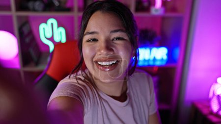 Téléchargez les photos : Une jeune femme hispanique heureuse qui prend un selfie dans une salle de jeux animée la nuit, avec des néons et une ambiance technologique. - en image libre de droit