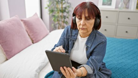 Ältere hispanische Frau nutzt Tablet mit Kopfhörer in gemütlicher Schlafzimmeratmosphäre und verkörpert den Einsatz moderner Technologie zu Hause.