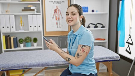 Photo for A young caucasian woman with tattoos smiles while holding a smartphone in a rehab clinic. - Royalty Free Image