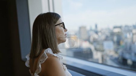 Schöne hispanische Geschäftsfrau mit Brille, fesselnder Blick auf die Stadt aus dem Fenster eines Wolkenkratzers in Japans urbaner Skyline