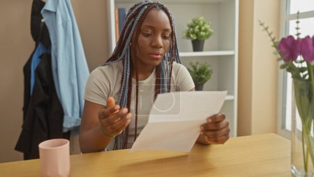 Téléchargez les photos : Une femme afro-américaine avec des tresses lit une lettre à une table à l'intérieur, symbolisant la contemplation dans un cadre domestique. - en image libre de droit