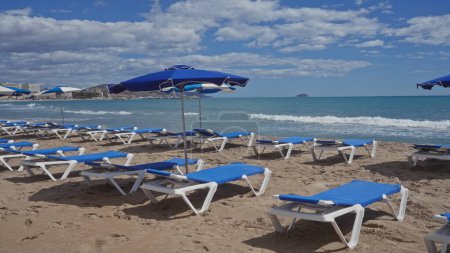 Photo for Empty blue sunbeds and umbrellas line a sandy beach with waves and clear skies hinting at a tranquil vacation setting. - Royalty Free Image