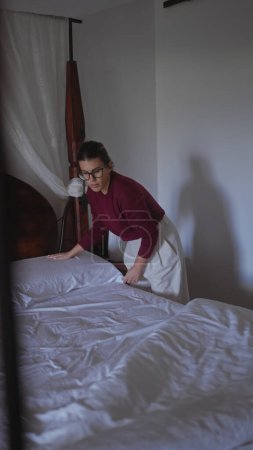 Téléchargez les photos : Une jeune femme concentrée arrange soigneusement des draps sur un lit à l'intérieur, mettant en valeur la vie quotidienne à la maison. - en image libre de droit