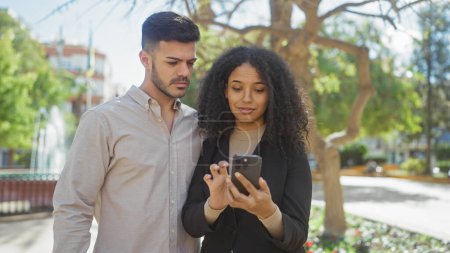 A man and woman stand together outdoors, focused on a smartphone in a sunlit park with greenery.