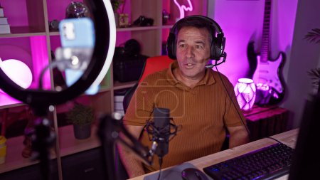 Téléchargez les photos : Un homme mature avec des écouteurs est assis dans une salle de jeux colorée la nuit, parlant dans un microphone avec une lumière annulaire et un ordinateur visible. - en image libre de droit