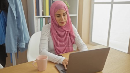 A focused woman wearing a pink hijab working on a laptop at home, representing modern muslim professional life.