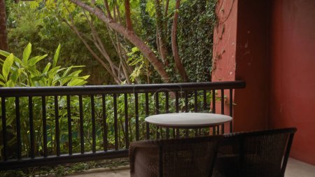 Foto de Tranquila escena de balcón con una mesa, sillas, plantas tropicales y una pared roja, lo que sugiere un relajante espacio al aire libre. - Imagen libre de derechos