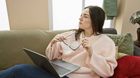 Thoughtful young woman with glasses sitting on a sofa in a cozy room, holding a laptop.