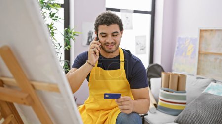 Smiling man holding credit card and talking on smartphone in art studio
