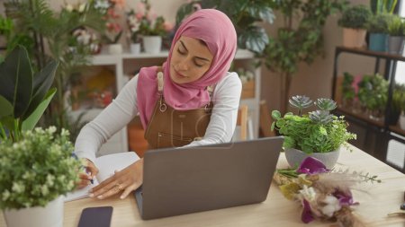 A focused woman in a hijab writes notes beside her laptop in a plant-filled home office
