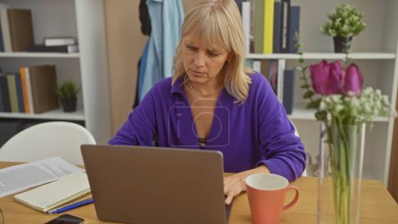 Mature woman working on laptop at home with coffee and notebook, displaying concentration and casual style.