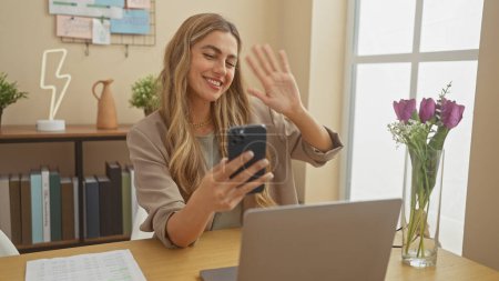Téléchargez les photos : Une jeune femme souriante s'engageant dans un appel vidéo à son bureau à domicile confortable, saluant à l'écran du smartphone. - en image libre de droit