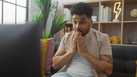 Foto de Un hombre afroamericano sorprendido con tatuajes reacciona emocionalmente mientras ve la televisión en su moderna sala de estar. - Imagen libre de derechos