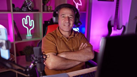 Middle-aged man with arms crossed smiles in a colorful, neon-lit gaming room at home.