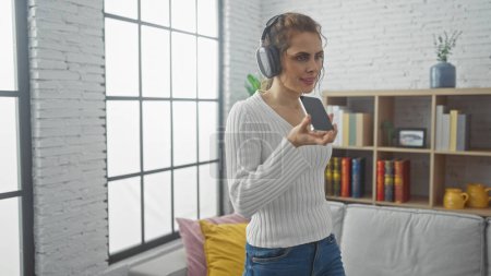 Photo for Young caucasian woman recording a voice message at home with headphones and smartphone, showing a modern interior background. - Royalty Free Image