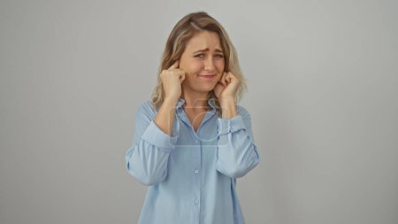 Mujer caucásica joven molesta en una camisa azul que cubre sus orejas contra una pared blanca, encarnando la frustración