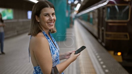 Foto de Mujer hispana alegre y hermosa inmersa en su smartphone, felizmente esperando su tren en la plataforma de la estación. - Imagen libre de derechos