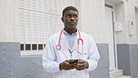 Un professionnel afro-américain en blouse de laboratoire avec un stéthoscope se tient devant un hôpital à l'aide d'un smartphone.