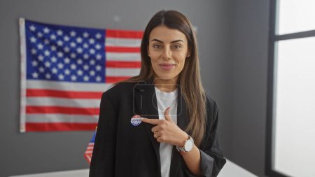 Una joven mujer hispana confiada señala su etiqueta de 'yo voté', mostrando compromiso cívico contra un telón de fondo de la bandera americana dentro de un ambiente interior.