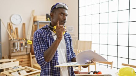 Hombre negro reflexivo en una camisa a cuadros planificación en un taller de carpintería con herramientas y madera alrededor