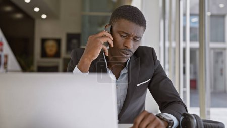 Foto de Hombre africano concentrado en traje de negocios trabajando y hablando por teléfono en un entorno de oficina. - Imagen libre de derechos