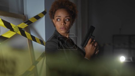 Téléchargez les photos : Femme noire concentrée tenant une arme sur une scène de crime faiblement éclairée avec ruban adhésif - en image libre de droit