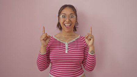 Excited young hispanic woman with glasses pointing up, isolated against a pink wall, wearing a striped shirt.