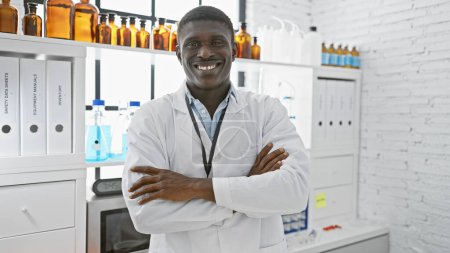 Téléchargez les photos : Homme africain confiant en blouse blanche debout avec les bras croisés dans un laboratoire lumineux - en image libre de droit