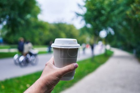 Téléchargez les photos : Main d'homme tenant une tasse de café dans un parc avec des piétons et des cyclistes flous en arrière-plan - en image libre de droit