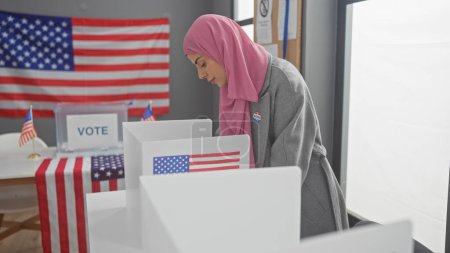 Una joven en un hiyab se involucra en la democracia americana, votando en una cabina adornada con la bandera de los Estados Unidos.