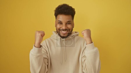 Un hombre afroamericano sonriente en una sudadera con capucha beige celebra contra un fondo amarillo.
