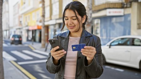 Téléchargez les photos : Young hispanic woman using a smartphone and holding a credit card on a city street. - en image libre de droit
