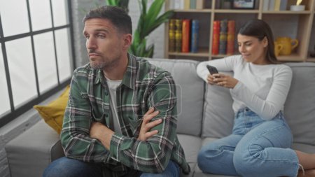 Upset man and woman using smartphone separately, highlighting relationship issues in a living room setting.