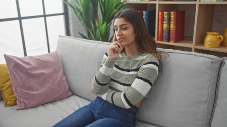 Téléchargez les photos : Thoughtful young hispanic woman relaxing on sofa at home, showcasing casual style and modern interior. - en image libre de droit