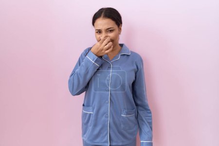 Foto de Mujer árabe joven usando pijama azul oliendo algo apestoso y asqueroso, olor intolerable, conteniendo la respiración con los dedos en la nariz. mal olor - Imagen libre de derechos