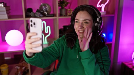 Téléchargez les photos : Une jeune femme souriante avec des écouteurs prend un selfie dans une salle de jeux vibrante et éclairée au néon la nuit. - en image libre de droit
