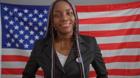 Foto de Mujer afroamericana confiada con trenzas usando una chaqueta posa en una oficina con la bandera de EE.UU.. - Imagen libre de derechos