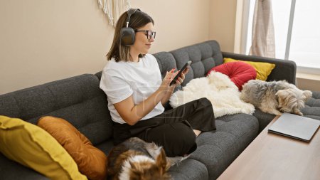 Téléchargez les photos : Femme hispanique se détendre avec smartphone et écouteurs sur le canapé entouré de chiens dans le salon confortable. - en image libre de droit