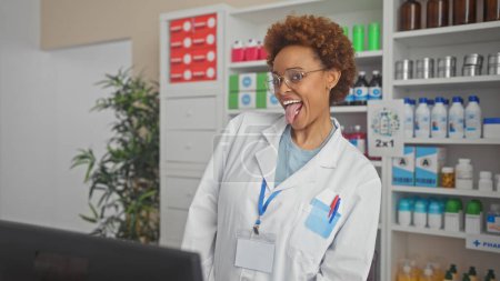 Photo for A cheerful african american woman pharmacist sticks out her tongue playfully in a well-stocked drugstore - Royalty Free Image