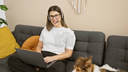 A cheerful young hispanic woman in glasses and headset uses a laptop in a cozy living room with her cute dog by her side.