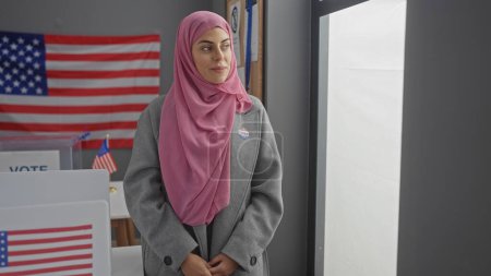 Una joven vistiendo un hijab se pone de pie con una expresión orgullosa en un centro de votación americano, mostrando una pegatina de 'yo voté'.