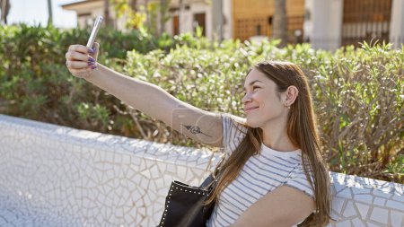 Téléchargez les photos : Femme souriante prenant selfie dans un parc ensoleillé avec verdure et banc - en image libre de droit