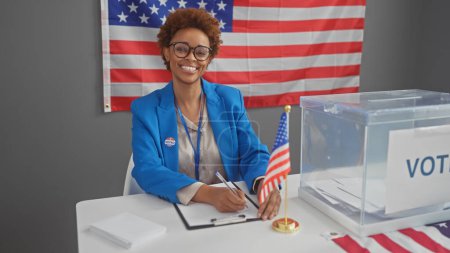Foto de Una sonriente mujer afroamericana en una chaqueta azul tomando notas en un centro electoral interior de Estados Unidos con una bandera americana. - Imagen libre de derechos