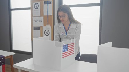 Foto de Una joven hispana observa una boleta en un centro de votación americano, adornado con banderas. - Imagen libre de derechos