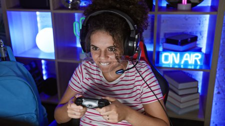 Smiling hispanic woman with curly hair gaming at night in a dark room illuminated by neon lights.