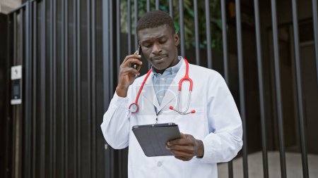 Photo for African american doctor outdoors in urban setting talking on phone while holding tablet - Royalty Free Image