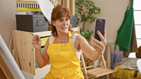 Photo for Middle-aged woman in a yellow apron taking a selfie with a smartphone in a creative studio space with art supplies. - Royalty Free Image