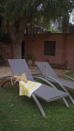 Foto de Un entorno tranquilo al aire libre con dos tumbonas, una toalla a rayas y un sombrero de paja en un contexto de una casa rústica y un árbol exuberante. - Imagen libre de derechos